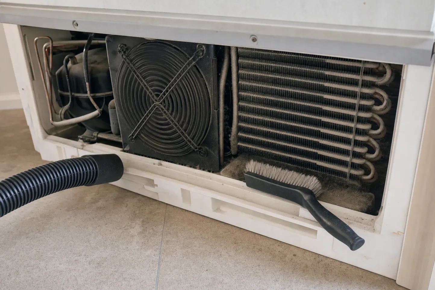Toe-kick grille removed from a low cabinet, showing the compressor, the condenser fan and a condenser matted with grey dust, a stiff brush and a vacuum hose at the floor