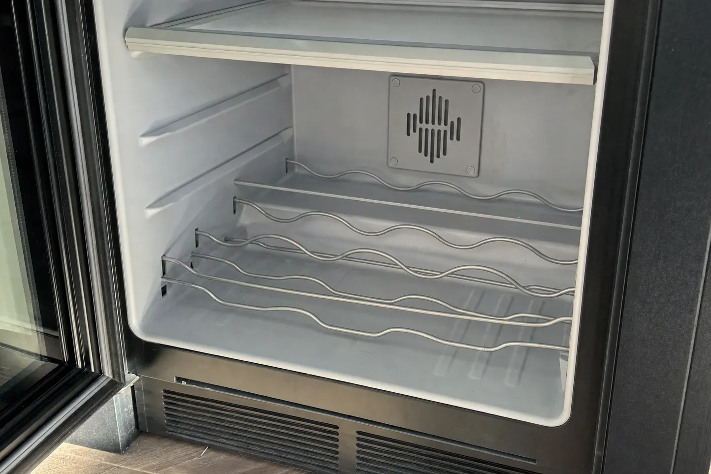 The inside of an empty glass-door undercounter cabinet: wave-form wire racks, a moulded air outlet high on the rear wall and a vent grille under the door